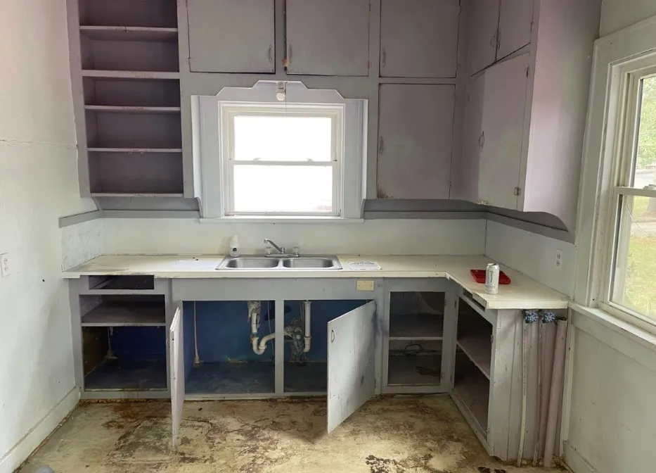 Kitchen before remodeling, with worn lilac-gray cabinets, missing cabinet doors, an old sink, and a damaged floor