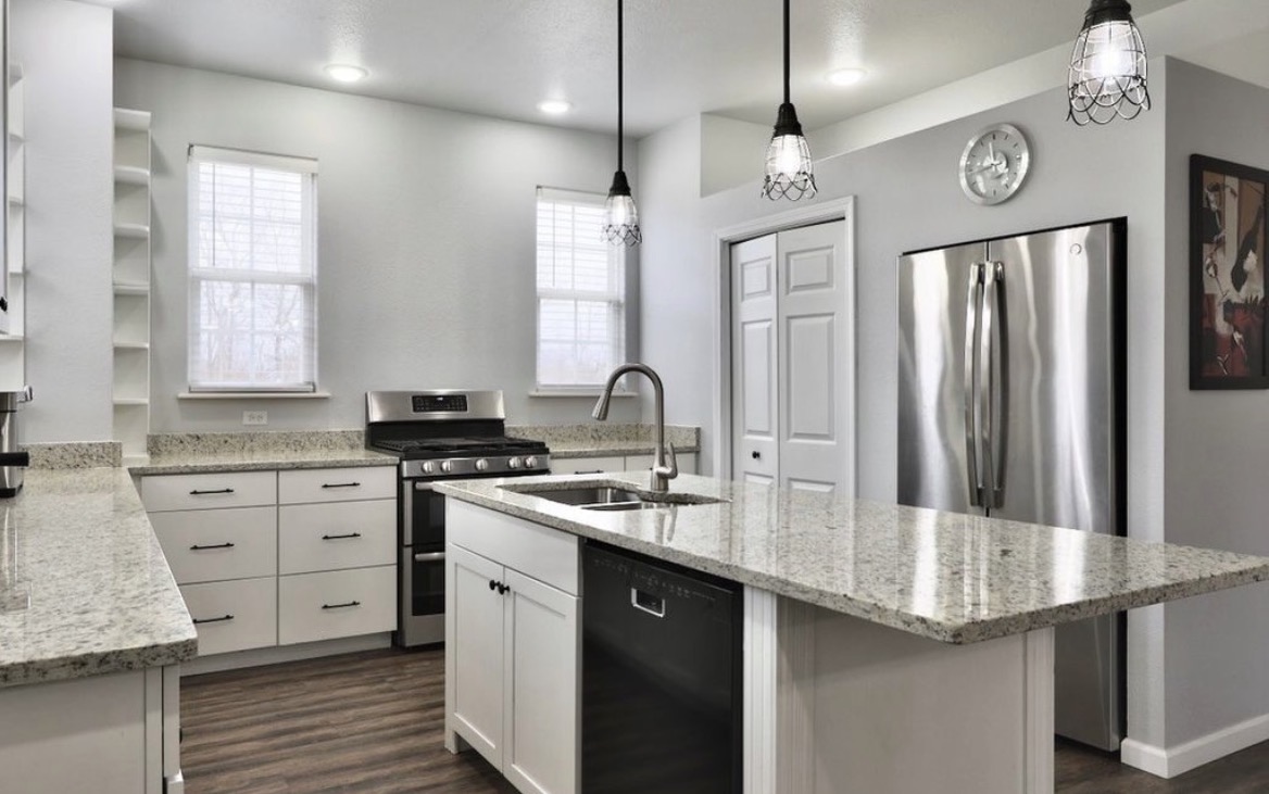 Angled view of a remodeled white kitchen with granite countertops, a center island, stainless appliances, and pendant lighting