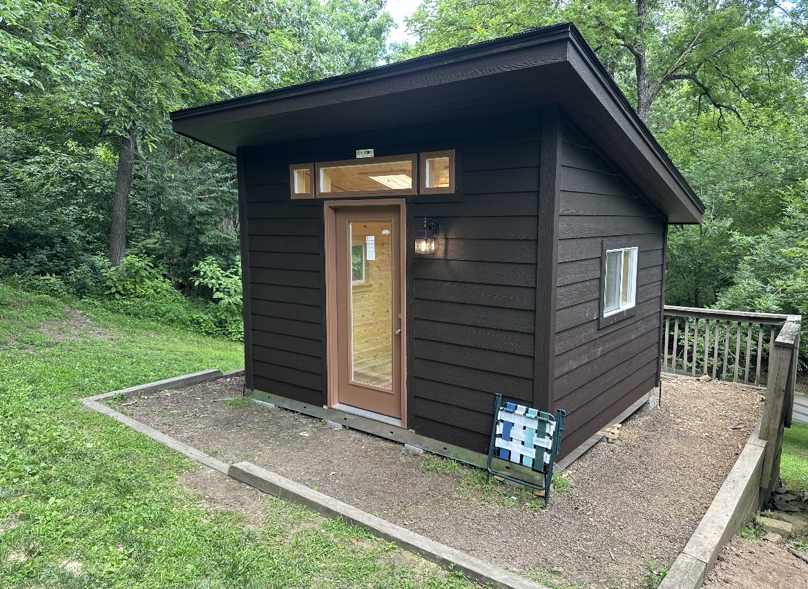 Dark brown modern lean-to shed installed and finished by Renew Home Solutions, with horizontal lap siding, a wood-and-glass entry door, and transom windows on a concrete pad in a wooded Springdale, AR yard