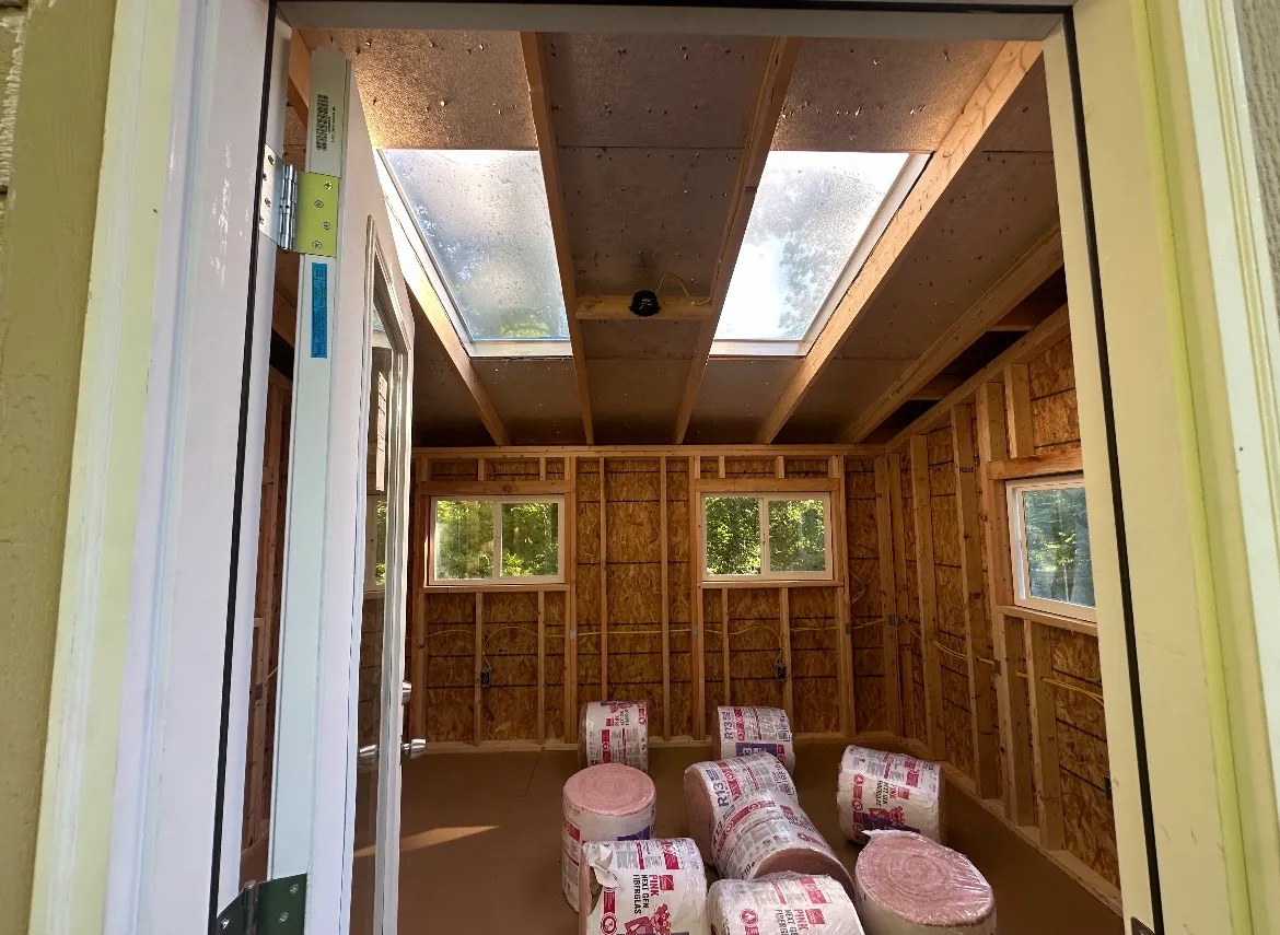 Interior of the shed during framing, with exposed studs, OSB sheathing, twin skylights, and rolls of insulation on the subfloor