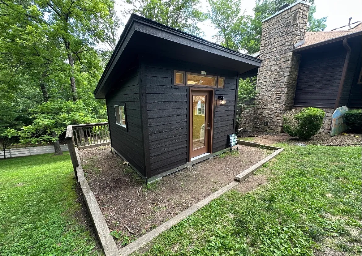 The same shed finished by Renew Home Solutions, with rich black-stained siding, trimmed transom windows and door, and a clean skirted base