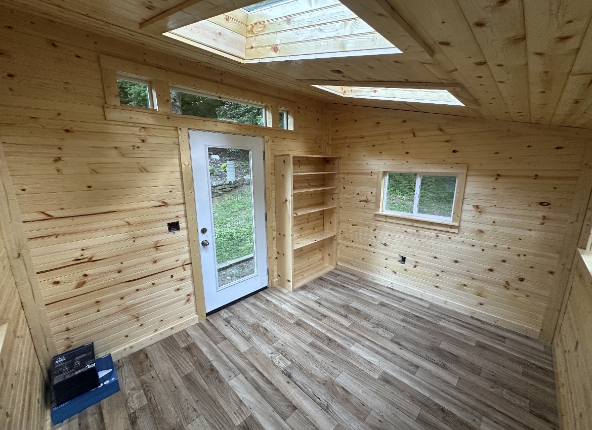 Finished shed interior with pine tongue-and-groove walls, skylights, built-in shelving, and vinyl plank flooring